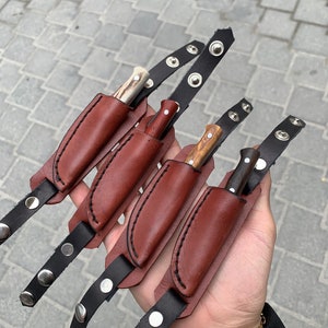 May include: Four small knives with wooden and bone handles, each in a brown leather sheath. The sheaths are attached to black leather straps with silver snap closures. The knives vary in handle color and design.