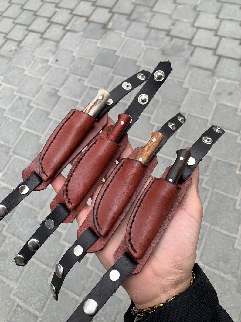 May include: Four small knives with wooden and bone handles, each in a brown leather sheath. The sheaths are attached to black leather straps with silver snap closures. The knives vary in handle color and design.