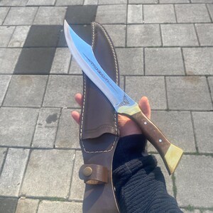 May include: A curved steel blade knife featuring an etched design on the metal, a wooden handle with brass accents, and two silver rivets. The knife is accompanied by a dark brown leather sheath with visible stitching and a brass snap closure.
