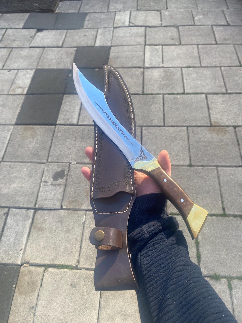 May include: A curved steel blade knife featuring an etched design on the metal, a wooden handle with brass accents, and two silver rivets. The knife is accompanied by a dark brown leather sheath with visible stitching and a brass snap closure.
