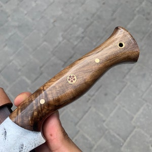 May include: Close-up of a handcrafted knife with a wooden handle. The handle is a rich brown color with decorative brass accents. The blade is partially visible, showing a textured, silver finish. The knife appears to be a hunting or utility knife.