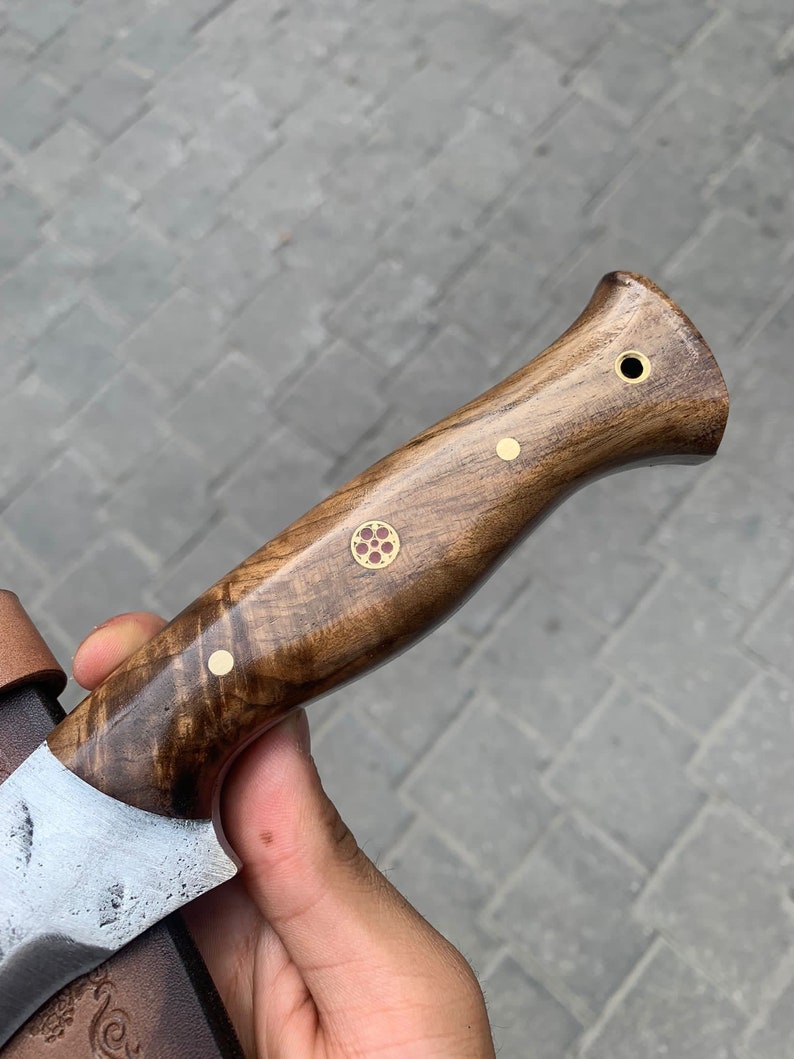 May include: Close-up of a handcrafted knife with a wooden handle. The handle is a rich brown color with decorative brass accents. The blade is partially visible, showing a textured, silver finish. The knife appears to be a hunting or utility knife.