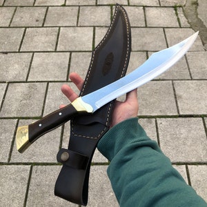 May include: A curved steel blade knife with a dark wood handle, brass bolster, and pommel. The knife is paired with a dark brown leather sheath featuring decorative stitching, a snap button closure, and an embossed pattern on the front surface.