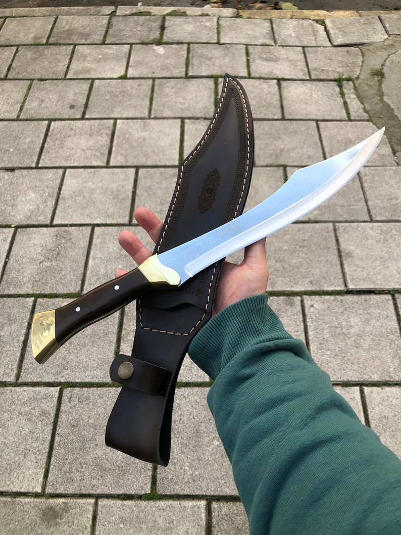 May include: A curved steel blade knife with a dark wood handle, brass bolster, and pommel. The knife is paired with a dark brown leather sheath featuring decorative stitching, a snap button closure, and an embossed pattern on the front surface.