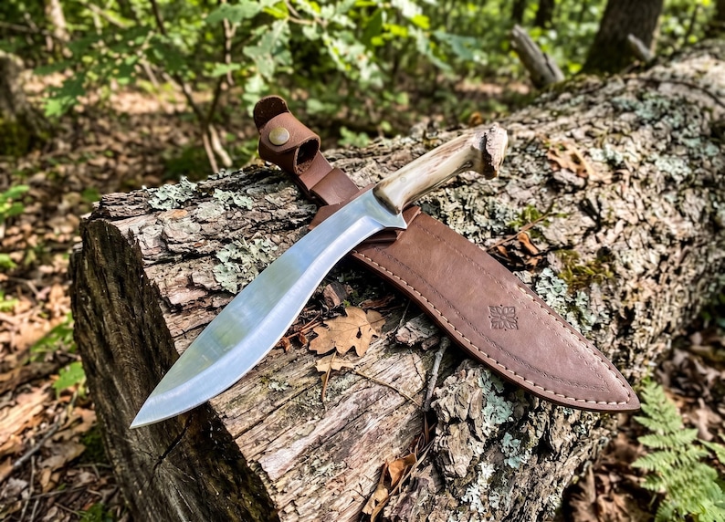 May include: A large, curved blade knife with a light-colored handle and a brown leather sheath. The knife is resting on a weathered log in a forest setting. The sheath has a decorative emblem.