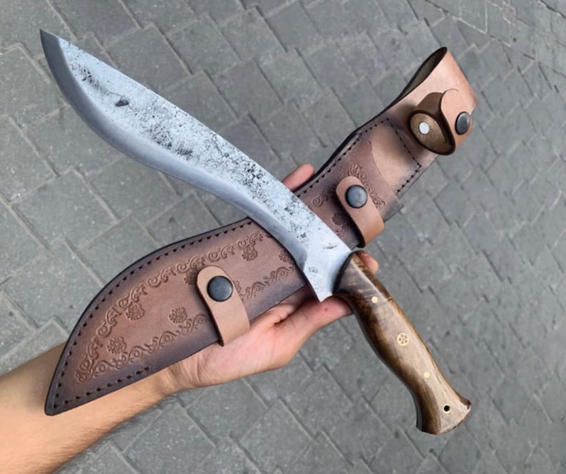 May include: A large, curved blade knife with a wooden handle and a leather sheath. The blade has a textured, silver finish. The sheath is brown leather with decorative floral embossing and a strap with a snap closure.
