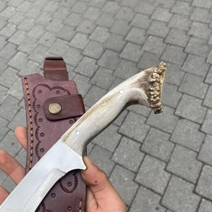 May include: A handmade hunting knife with a polished steel blade and a handle made of antler. The knife is paired with a brown leather sheath with decorative stitching and a snap closure. The blade is approximately 8 inches long.