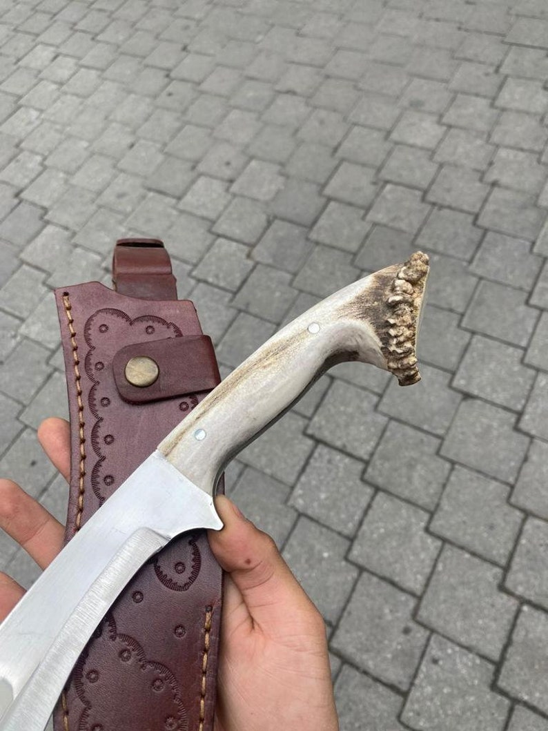 May include: A handmade hunting knife with a polished steel blade and a handle made of antler. The knife is paired with a brown leather sheath with decorative stitching and a snap closure. The blade is approximately 8 inches long.