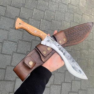 May include: A large, handmade knife with a light wood handle and a textured, silver-colored blade. The knife is paired with a brown leather sheath, featuring decorative embossing and secured with brass-colored snaps. The knife is approximately 12 inches long.