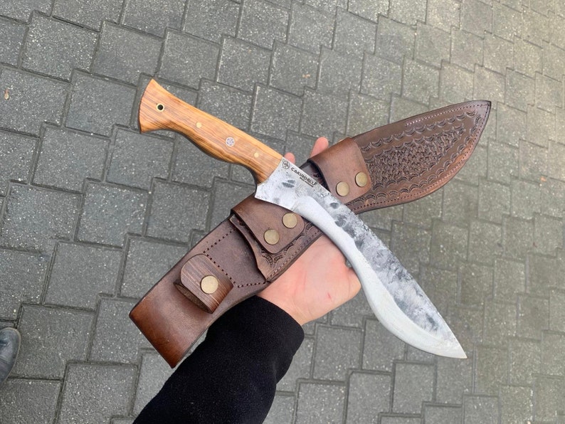May include: A large, handmade knife with a light wood handle and a textured, silver-colored blade. The knife is paired with a brown leather sheath, featuring decorative embossing and secured with brass-colored snaps. The knife is approximately 12 inches long.