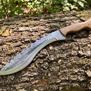May include: A large, curved blade knife with a wooden handle. The blade has a textured, gray finish and a sharp edge. The handle is a light brown color and is secured with small rivets. The knife is resting on a textured, brown log.