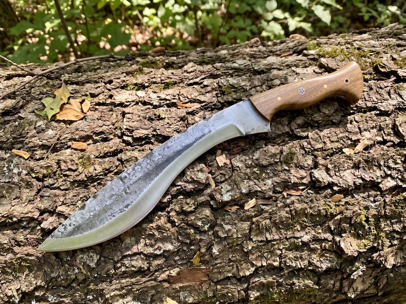 May include: A large, curved blade knife with a wooden handle. The blade has a textured, gray finish and a sharp edge. The handle is a light brown color and is secured with small rivets. The knife is resting on a textured, brown log.