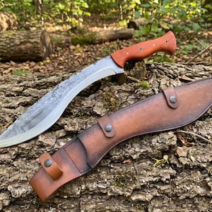 May include: A large, curved knife with a wooden handle and a leather sheath. The blade has a textured finish. The sheath is brown and has a decorative pattern. The knife and sheath are resting on a log.