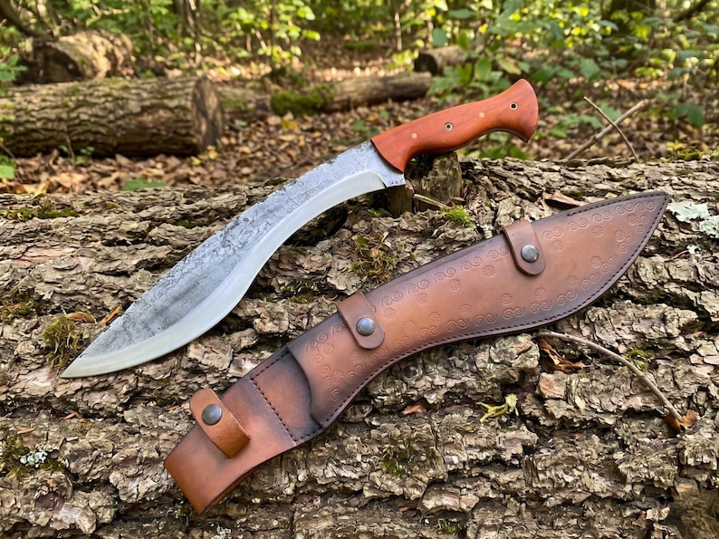 May include: A large, curved knife with a wooden handle and a leather sheath. The blade has a textured finish. The sheath is brown and has a decorative pattern. The knife and sheath are resting on a log.