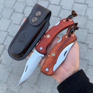 May include: Two pocket knives with wooden handles and silver blades, alongside a dark brown leather sheath. The knives have a classic design with brass accents. The sheath has two decorative metal buttons.
