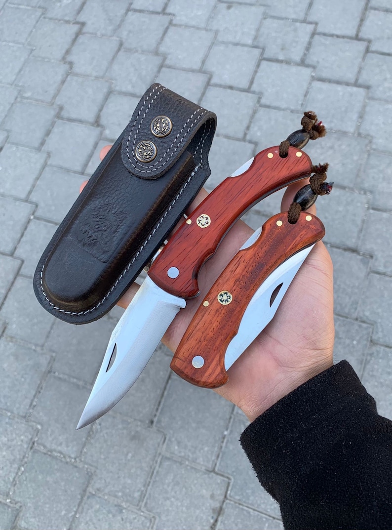 May include: Two pocket knives with wooden handles and silver blades, alongside a dark brown leather sheath. The knives have a classic design with brass accents. The sheath has two decorative metal buttons.