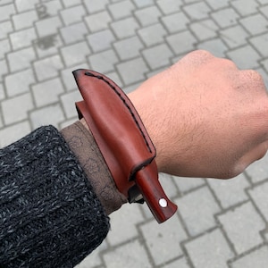 May include: A brown leather sheath with a small knife. The knife has a wooden handle with a white dot and a dark blade. The sheath is stitched with black thread.