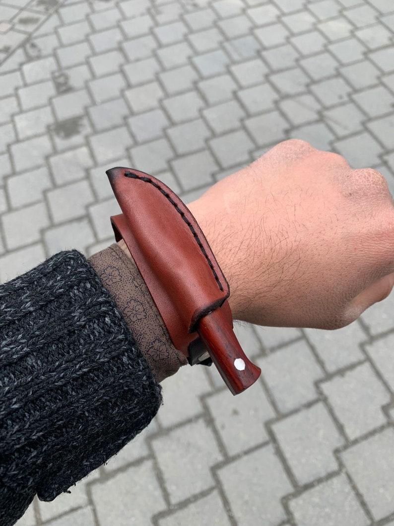 May include: A brown leather sheath with a small knife. The knife has a wooden handle with a white dot and a dark blade. The sheath is stitched with black thread.
