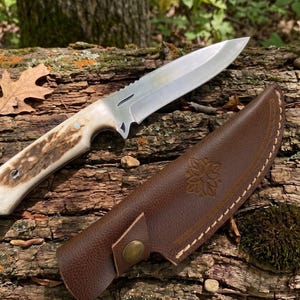May include: A hunting knife with a stainless steel blade and a handle with a natural antler design. The knife rests on a weathered log next to a brown leather sheath with a floral design.