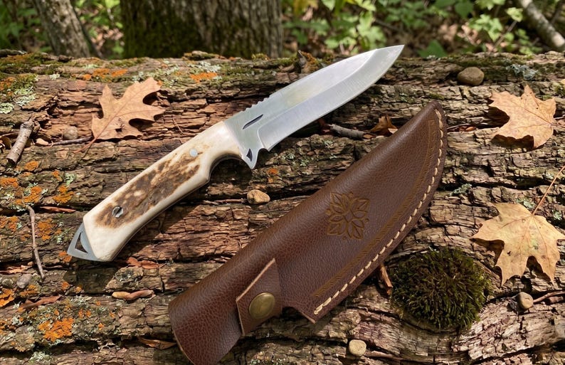 May include: A hunting knife with a stainless steel blade and a handle with a natural antler design. The knife rests on a weathered log next to a brown leather sheath with a floral design.