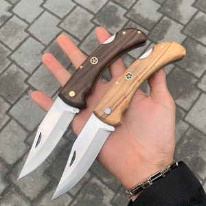 May include: Two folding pocket knives with wooden handles. One knife has a dark brown handle, the other a light brown handle. Both knives have silver blades and brass accents. The blades are open, displaying their sharp edges.