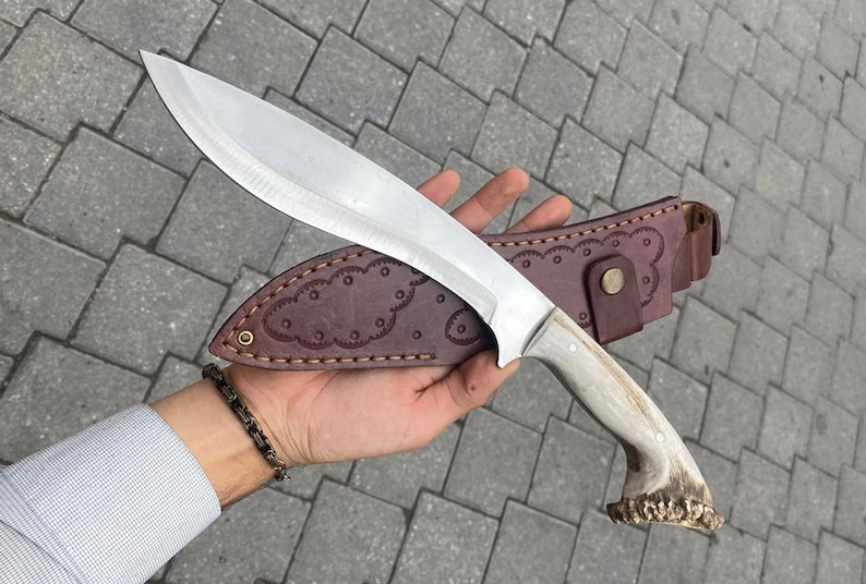 A handmade kukri machete held in a hand, featuring a deer antler handle with natural beige and brown tones, accented by two brass pins and a lanyard hole. The 20 cm stainless steel blade has a curved, polished finish typical of a kukri. A brown leather sheath with simple stitching lies beside the machete on a paved stone surface.
