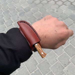 May include: A small, handcrafted knife with a wooden handle and a brown leather sheath. The knife has a silver-colored rivet on the handle. The sheath is stitched with black thread. The knife is designed for outdoor use.