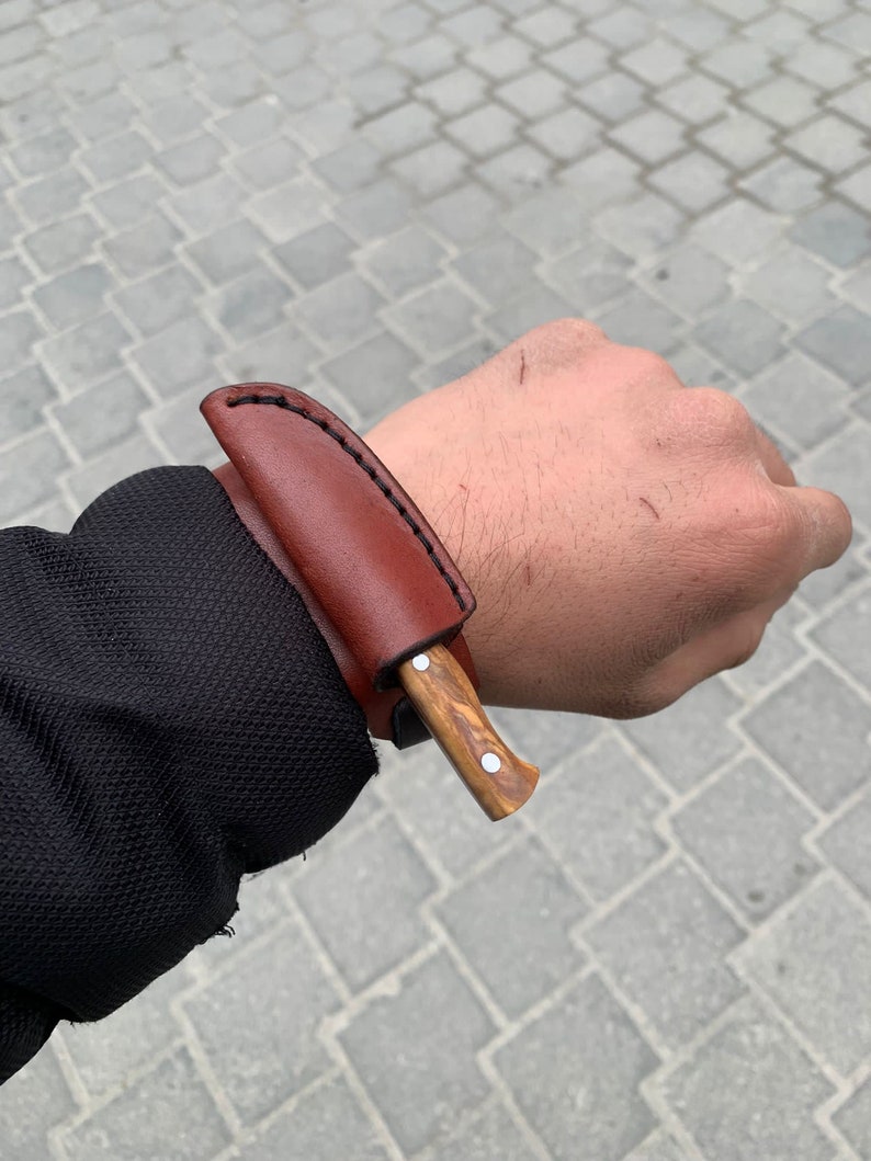 May include: A small, handcrafted knife with a wooden handle and a brown leather sheath. The knife has a silver-colored rivet on the handle. The sheath is stitched with black thread. The knife is designed for outdoor use.