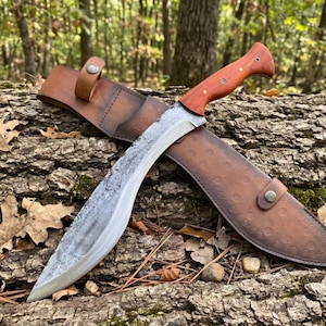 May include: A large, curved knife with a wooden handle and a leather sheath. The blade has a textured finish. The sheath is brown with decorative patterns and a snap closure. The knife is resting on a log.
