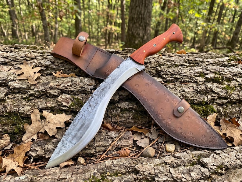 May include: A large, curved knife with a wooden handle and a leather sheath. The blade has a textured finish. The sheath is brown with decorative patterns and a snap closure. The knife is resting on a log.