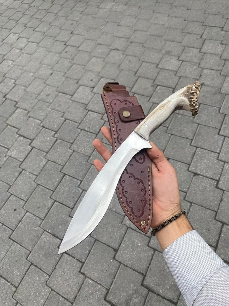May include: A large, curved hunting knife with a polished steel blade and a light-colored handle. The handle has a textured, antler-like appearance. The knife is in a brown leather sheath with decorative stitching and a snap closure. A bracelet is visible on the wrist.