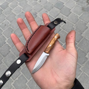 May include: A small, handcrafted knife with a wooden handle and a silver blade, accompanied by a brown leather sheath and a black leather strap with silver rivets. The knife is designed for outdoor use.