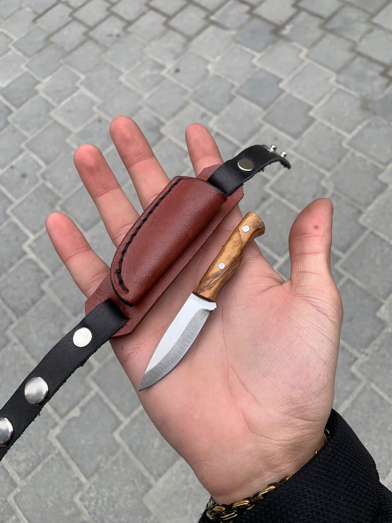 May include: A small, handcrafted knife with a wooden handle and a silver blade, accompanied by a brown leather sheath and a black leather strap with silver rivets. The knife is designed for outdoor use.