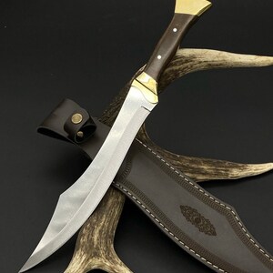 May include: A fixed-blade knife with a silver-toned steel blade, a dark brown wooden handle with two silver pins, and a brass bolster and pommel. The knife is accompanied by a dark brown leather sheath featuring decorative stitching and a brass snap.