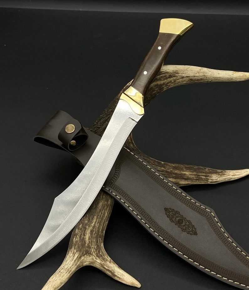 May include: A fixed-blade knife with a silver-toned steel blade, a dark brown wooden handle with two silver pins, and a brass bolster and pommel. The knife is accompanied by a dark brown leather sheath featuring decorative stitching and a brass snap.