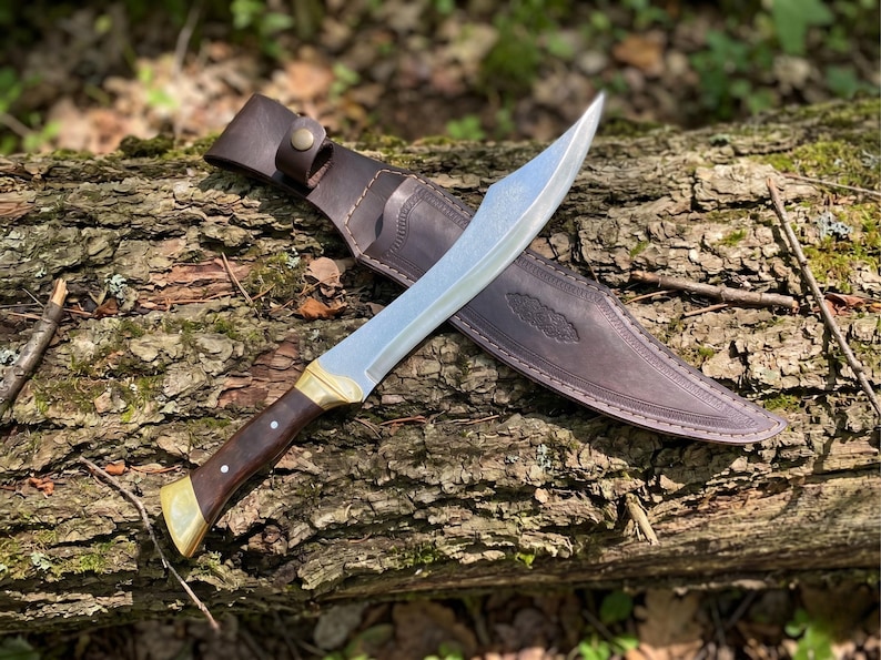 May include: A fixed-blade knife with a curved silver steel blade, a dark brown wooden handle with two silver rivets, and brass bolsters. The knife is accompanied by a dark brown leather sheath featuring decorative stitching and a snap closure.