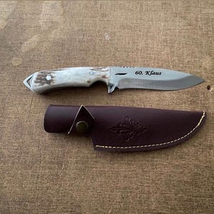 May include: A hunting knife with a silver blade and a light-coloured handle, resting on a wooden surface. The blade has the inscription "60. Klaus". A brown leather sheath is also present.