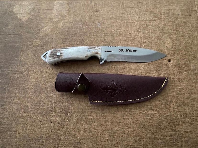 May include: A hunting knife with a silver blade and a light-coloured handle, resting on a wooden surface. The blade has the inscription "60. Klaus". A brown leather sheath is also present.