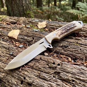 May include: A hunting knife with a polished steel blade and a handle made of bone and textured brown material. The knife is lying on a weathered log in a forest setting. The blade is approximately 20 cm long.