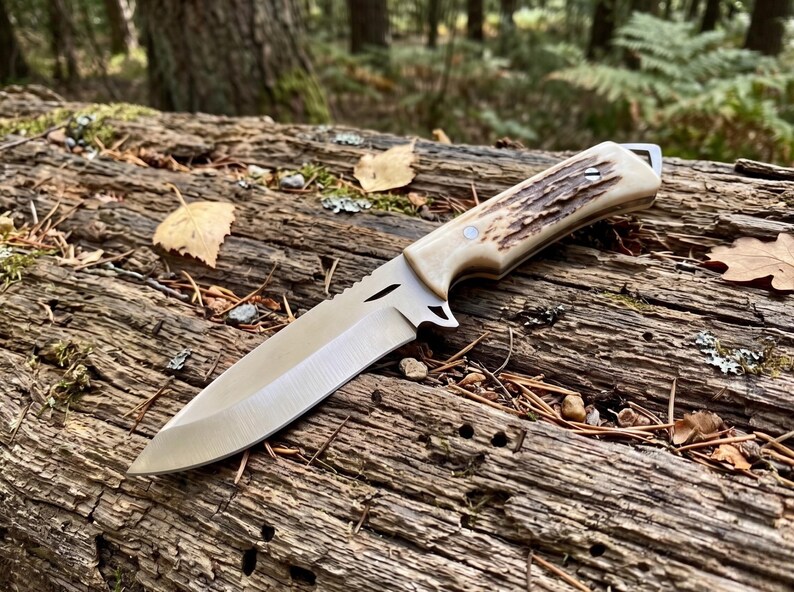 May include: A hunting knife with a polished steel blade and a handle made of bone and textured brown material. The knife is lying on a weathered log in a forest setting. The blade is approximately 20 cm long.