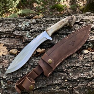 May include: A large, curved hunting knife with a polished steel blade and a light-colored antler handle. The knife rests on a textured log with its brown leather sheath. The sheath has a decorative emblem and a belt loop.