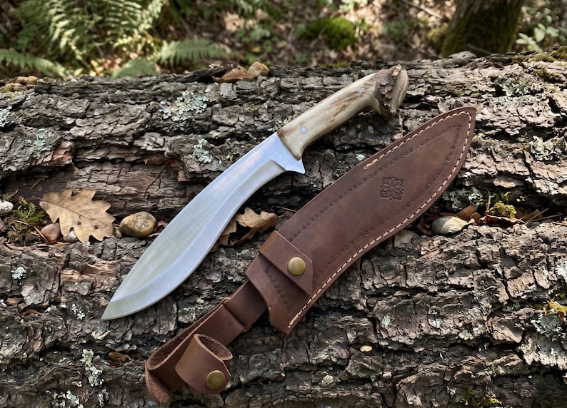 May include: A large, curved hunting knife with a polished steel blade and a light-colored antler handle. The knife rests on a textured log with its brown leather sheath. The sheath has a decorative emblem and a belt loop.