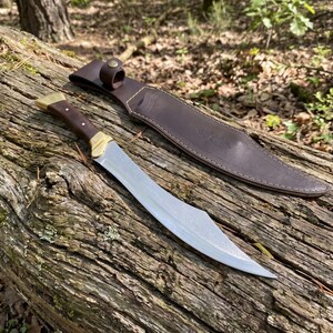 May include: A fixed-blade knife with a curved steel blade, a dark wood handle, and brass accents, accompanied by a brown leather sheath featuring a snap closure and visible stitching along the edges, designed for outdoor utility and carry.