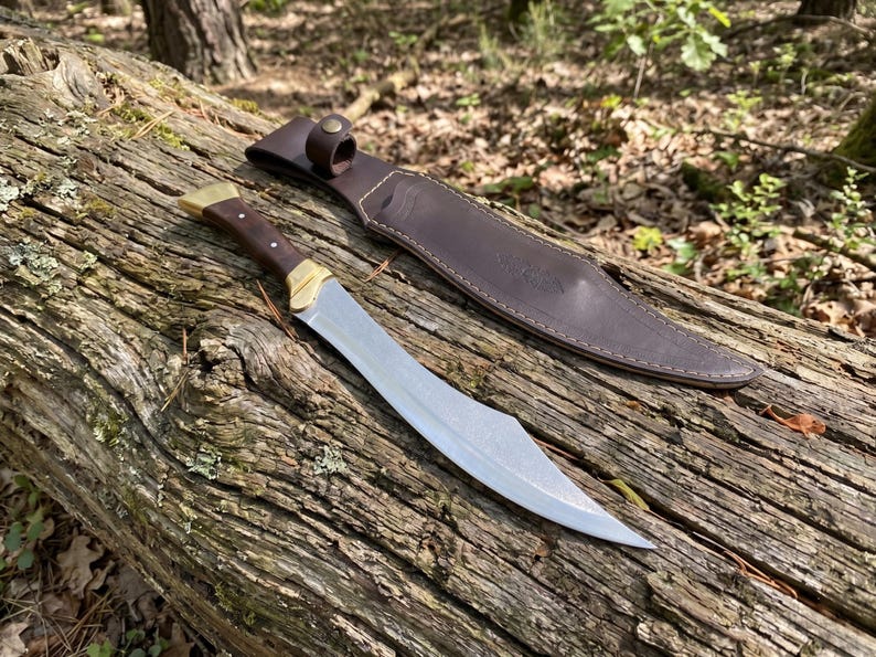 May include: A fixed-blade knife with a curved steel blade, a dark wood handle, and brass accents, accompanied by a brown leather sheath featuring a snap closure and visible stitching along the edges, designed for outdoor utility and carry.