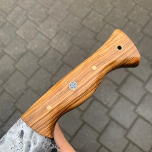 May include: A handcrafted knife with a wooden handle featuring a natural wood grain pattern. The handle is adorned with brass rivets and a decorative blue and gold inlay. The blade has a textured, hammered finish.