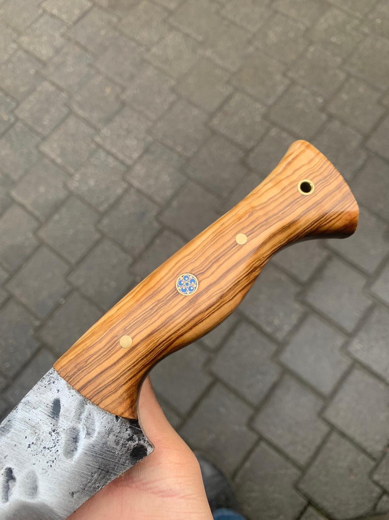 May include: A handcrafted knife with a wooden handle featuring a natural wood grain pattern. The handle is adorned with brass rivets and a decorative blue and gold inlay. The blade has a textured, hammered finish.