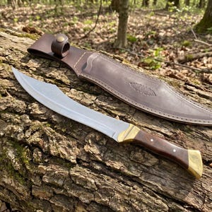 May include: A curved steel blade knife with a dark wood handle, brass bolster, and brass pommel. The knife is accompanied by a brown leather sheath featuring a snap button closure and decorative embossed detailing along the surface of the leather.