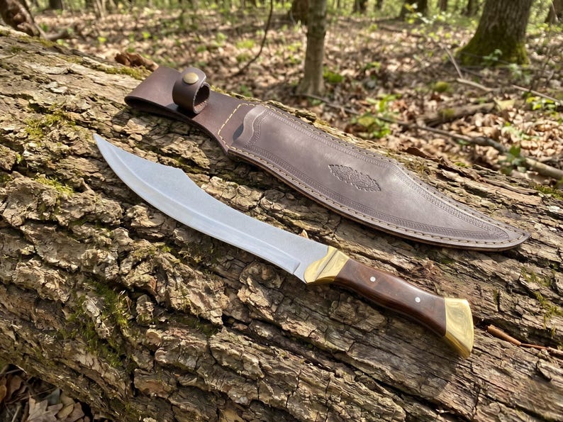 May include: A curved steel blade knife with a dark wood handle, brass bolster, and brass pommel. The knife is accompanied by a brown leather sheath featuring a snap button closure and decorative embossed detailing along the surface of the leather.