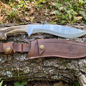 May include: A large, curved blade knife with a light-colored handle and a brown leather sheath. The blade is silver and the handle appears to be made of antler. The sheath has a decorative floral design and a belt loop. The knife and sheath are resting on a log.