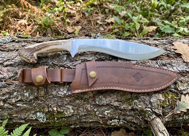 May include: A large, curved blade knife with a light-colored handle and a brown leather sheath. The blade is silver and the handle appears to be made of antler. The sheath has a decorative floral design and a belt loop. The knife and sheath are resting on a log.